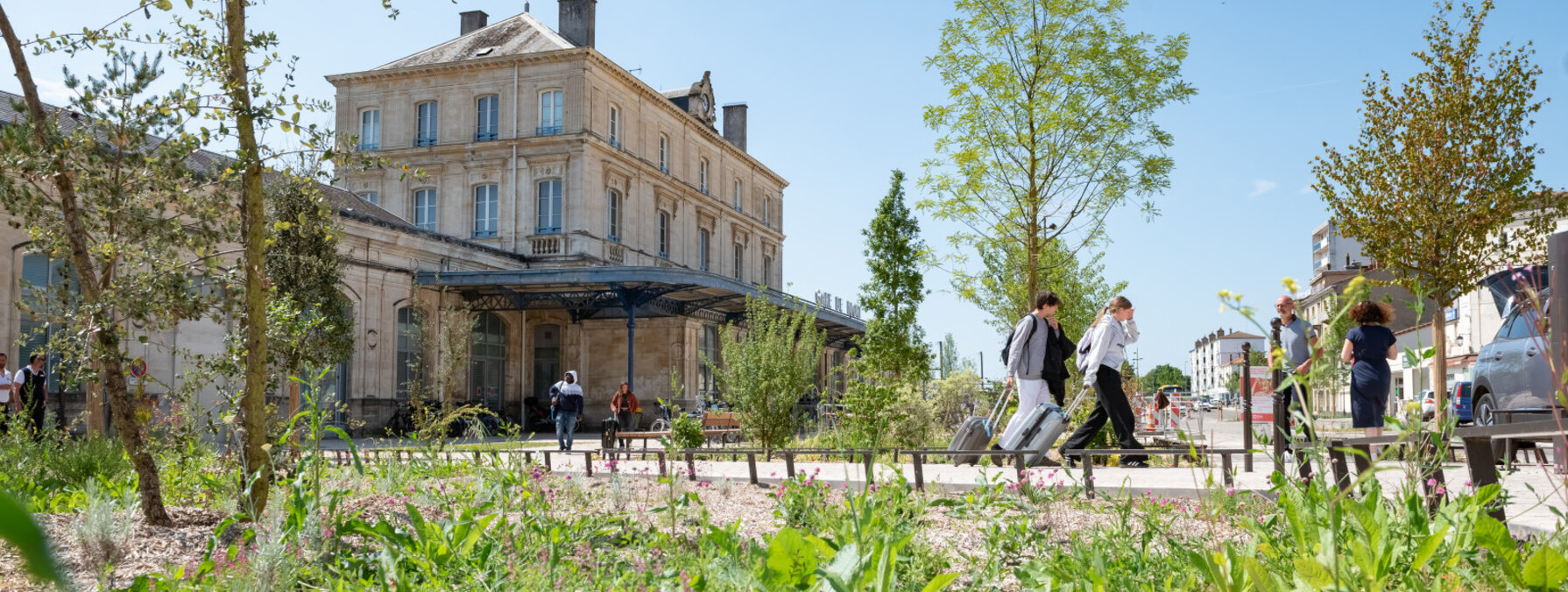 Esplanade réaménagée devant la gare de Niort avec jeunes arbres et plantations, des voyageurs avec bagages circulent