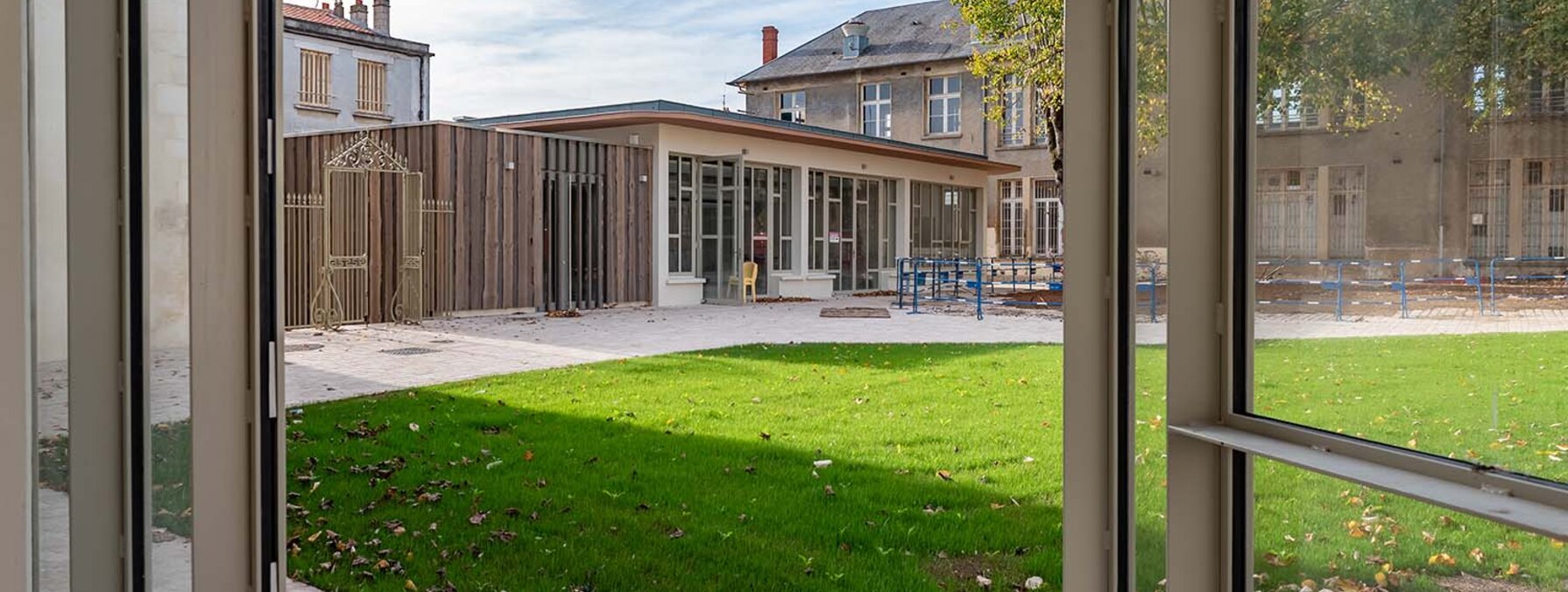 Chantier en fin de réhabilitation : pelouse et pavillon en bois neuf encadrés de bâtiments anciens, vus depuis une baie vitrée ouverte.