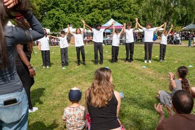 Groupe de jeunes bras levés et mains jointes lors d'une animation associative en plein air, devant un public réuni dans un parc.