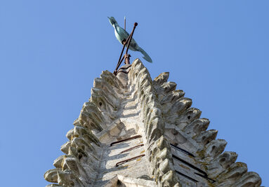 Flèche de pierre sculptée en écailles gothiques, surmontée d'un oiseau en cuivre patiné vert, sous un ciel bleu dégagé