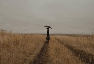 une femme dans un champ. Elle s'abrite sous un parapluie et tient une sacoche à la main.