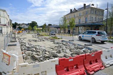 Dalles de béton brisées au sol et barrières de sécurité rouge et blanc délimitant le chantier, façade de la gare en arrière-plan.