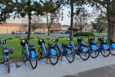 Sept vélos bleus en libre-service alignés en rang sur un emplacement balisé, avec une borne de déverrouillage et des espaces verts en arrière-plan